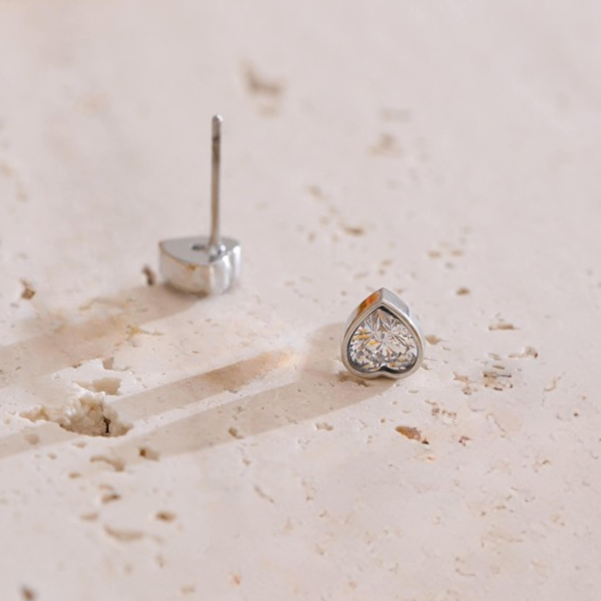 Heart-shaped silver earring with clear gemstone on a beige surface